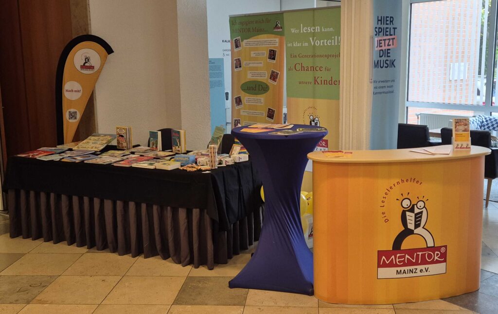 Foto vom Stand des Vereins aus der Büchermesse 2025 mit einer Theke, einem Tisch und den Büchern aus der Mentorkiste und weiteren Werbematerialien